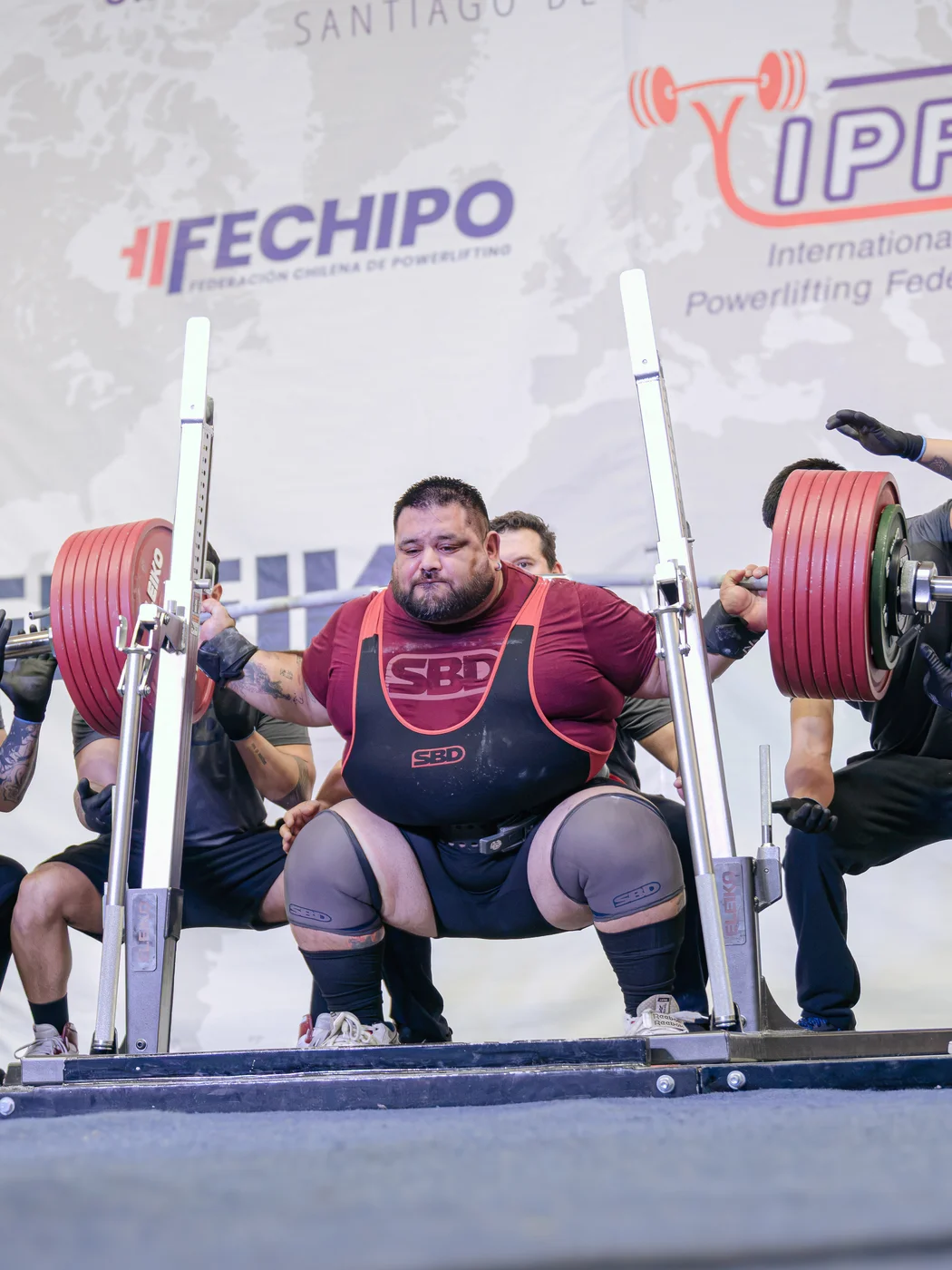 Atleta en el fondo de una sentadilla pesada, rodeado de ayudantes